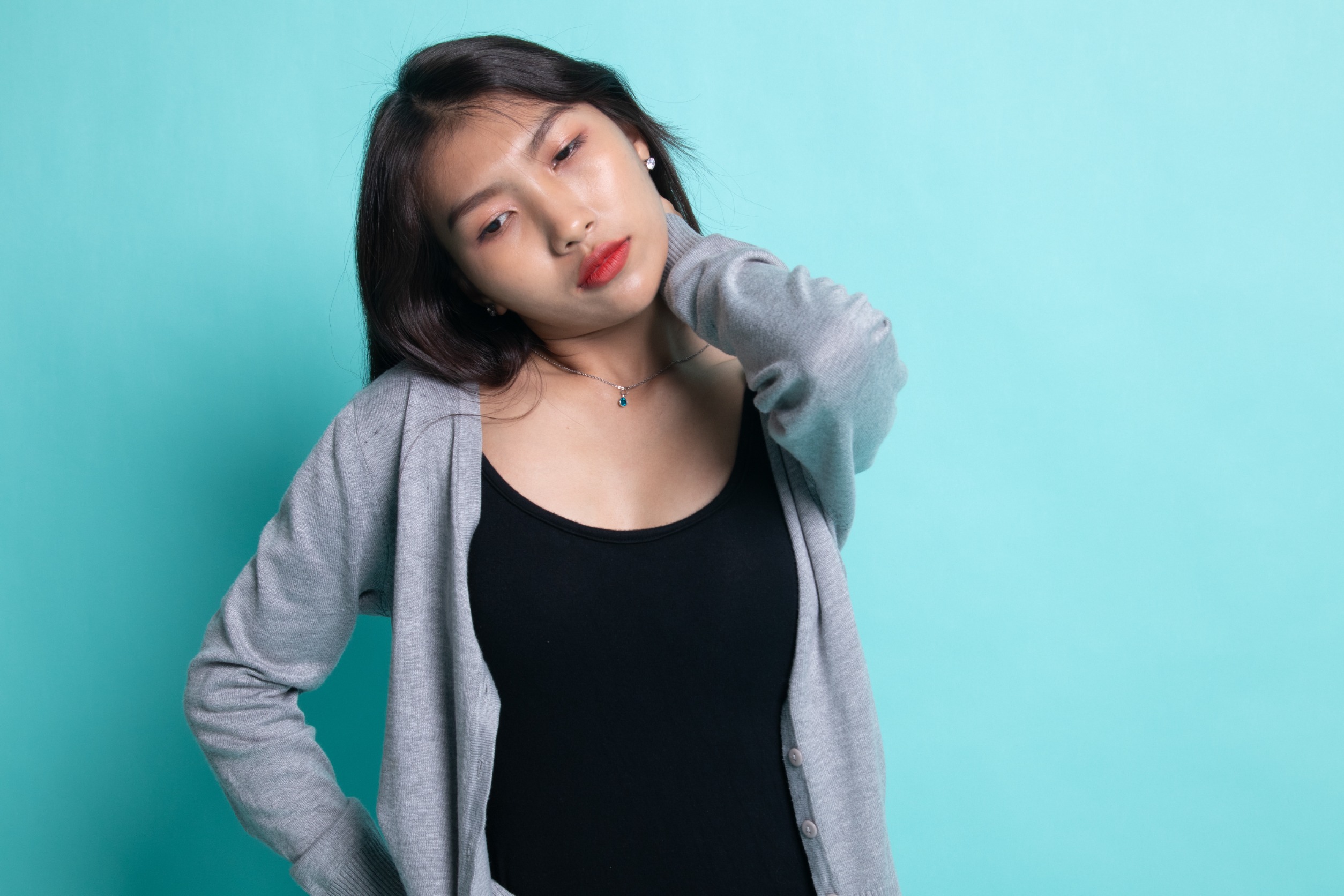 Une jeune femme asiatique a des douleurs au cou (cervicalgie) sur fond cyan.